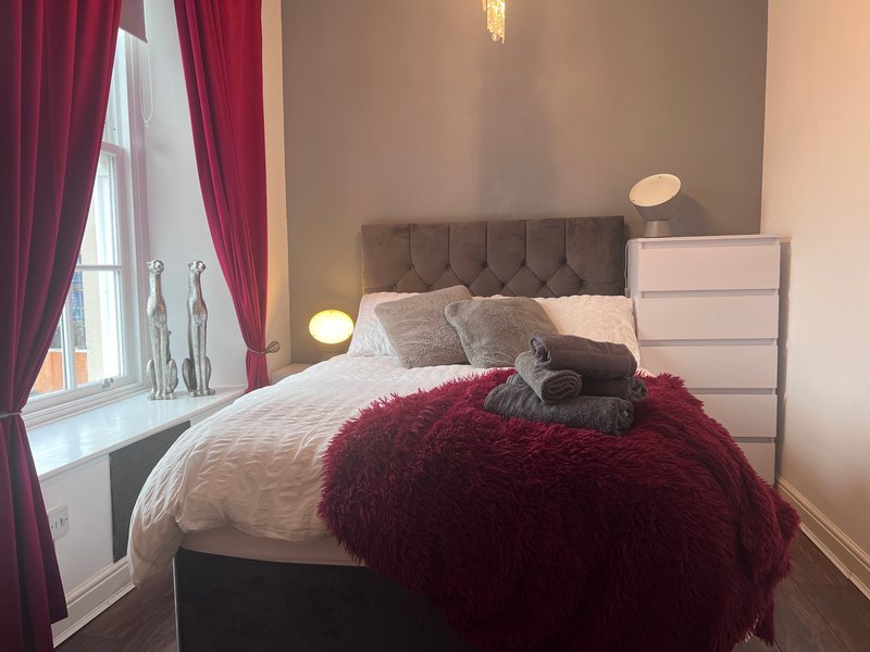 Modern bedroom with double bed, burgundy throw, grey headboard, white bedside table and red curtains