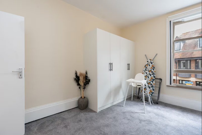 Bright bedroom with white wardrobe, grey carpet, window overlooking street, and minimalist decor