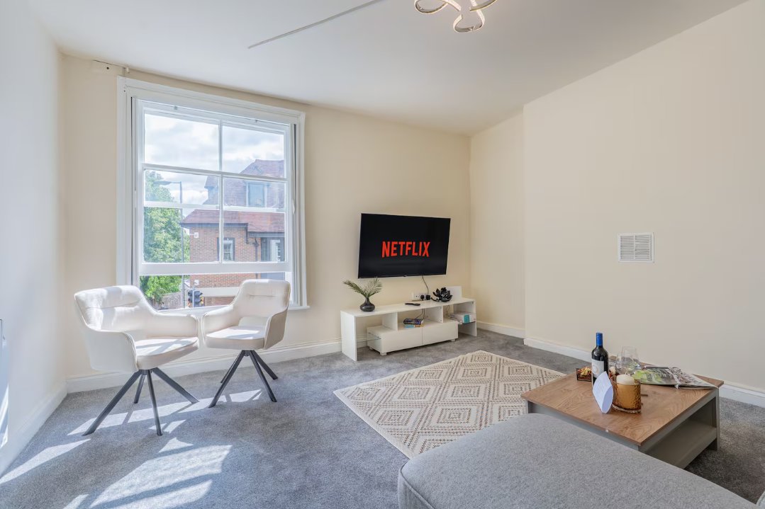 Modern living room with white chairs, TV on stand, large window and grey carpet flooring