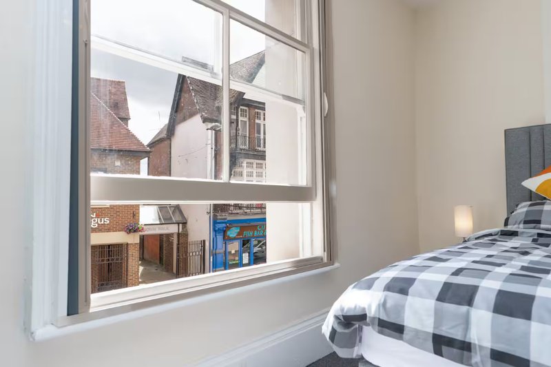 Bedroom with double bed, checkered duvet, window overlooking brick buildings and residential street