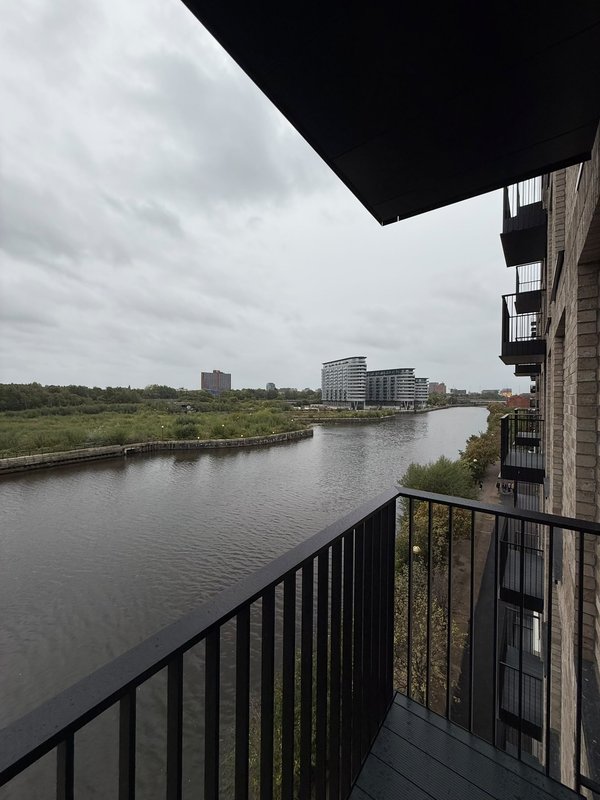 Balcony view over river with modern buildings and green banks under cloudy sky