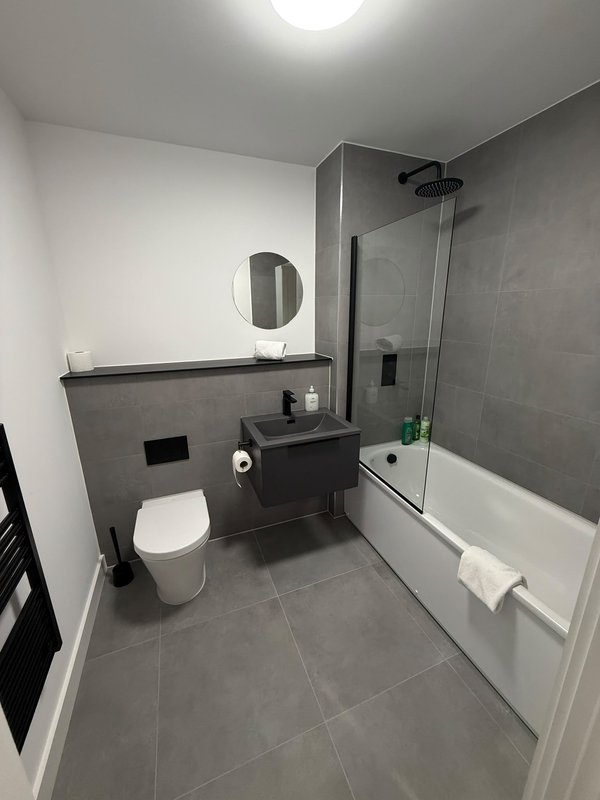 Modern bathroom with grey tile floor, white toilet, dark vanity, round mirror and glass shower enclosure