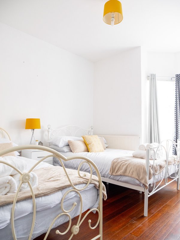 Bright bedroom with two white metal-frame beds, yellow lamp, and wooden flooring