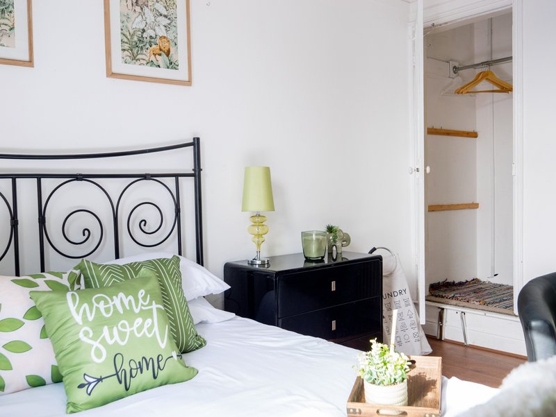 Bright bedroom with decorative wrought-iron bed frame, green patterned cushion, black bedside table, and open wardrobe