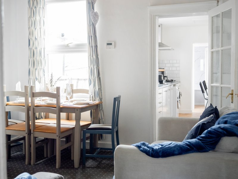 Open-plan living area with dining table, chair, sofa with blue cushions, and kitchen visible through doorway