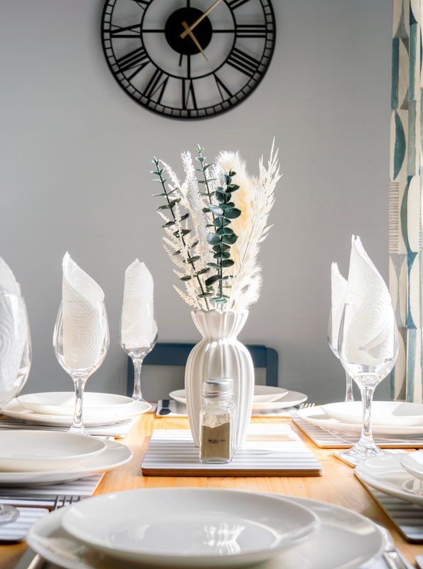Formal dining table with place settings, decorative vase with dried flowers, and wall clock