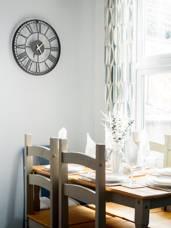 Bright dining area with wooden chairs, table, wall clock, and patterned curtains by window