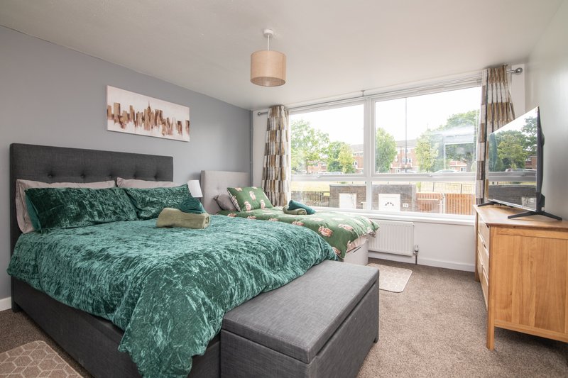 Bright bedroom with green patterned bedding, dark headboard, wooden dresser and large windows overlooking garden