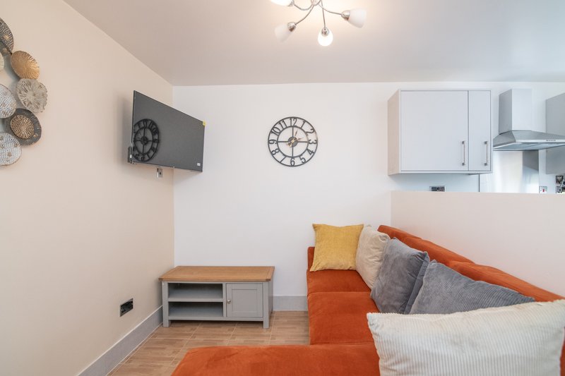 Modern living room with orange sofa, wall-mounted TV, kitchen area, and wall clock