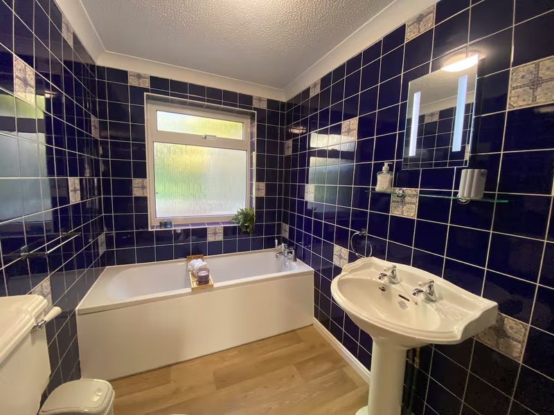 Bathroom with white bathtub, pedestal sink, dark tile walls and wooden floor