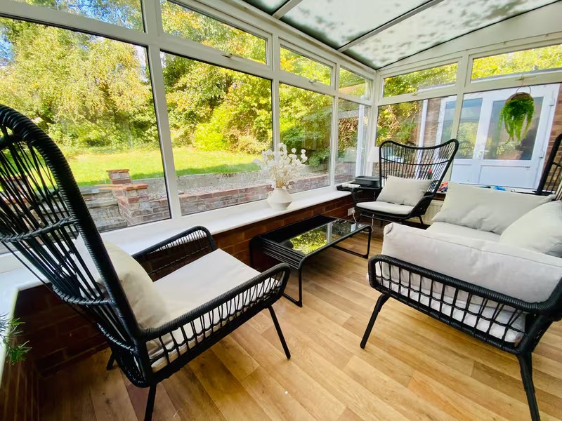 Bright conservatory with black wicker chairs, wooden table, and garden views through glass roof