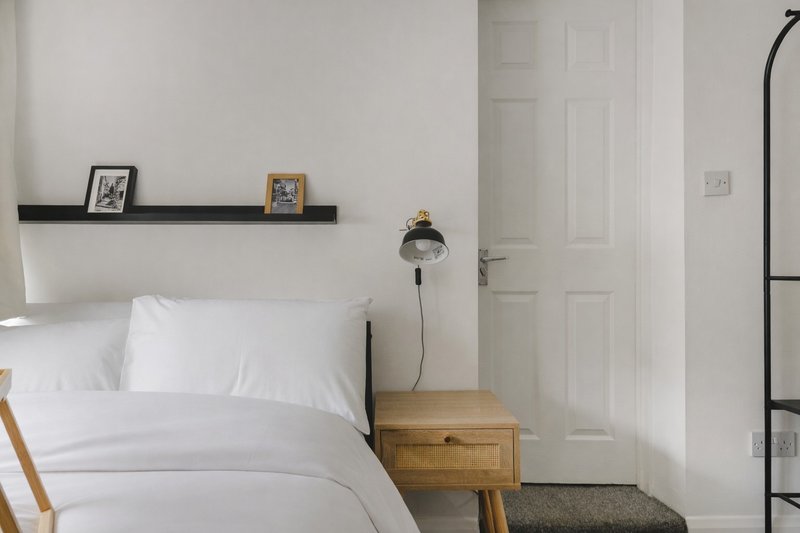 Minimalist bedroom with white bedding, wooden nightstand, floating shelf with framed photos, and pendant lamp
