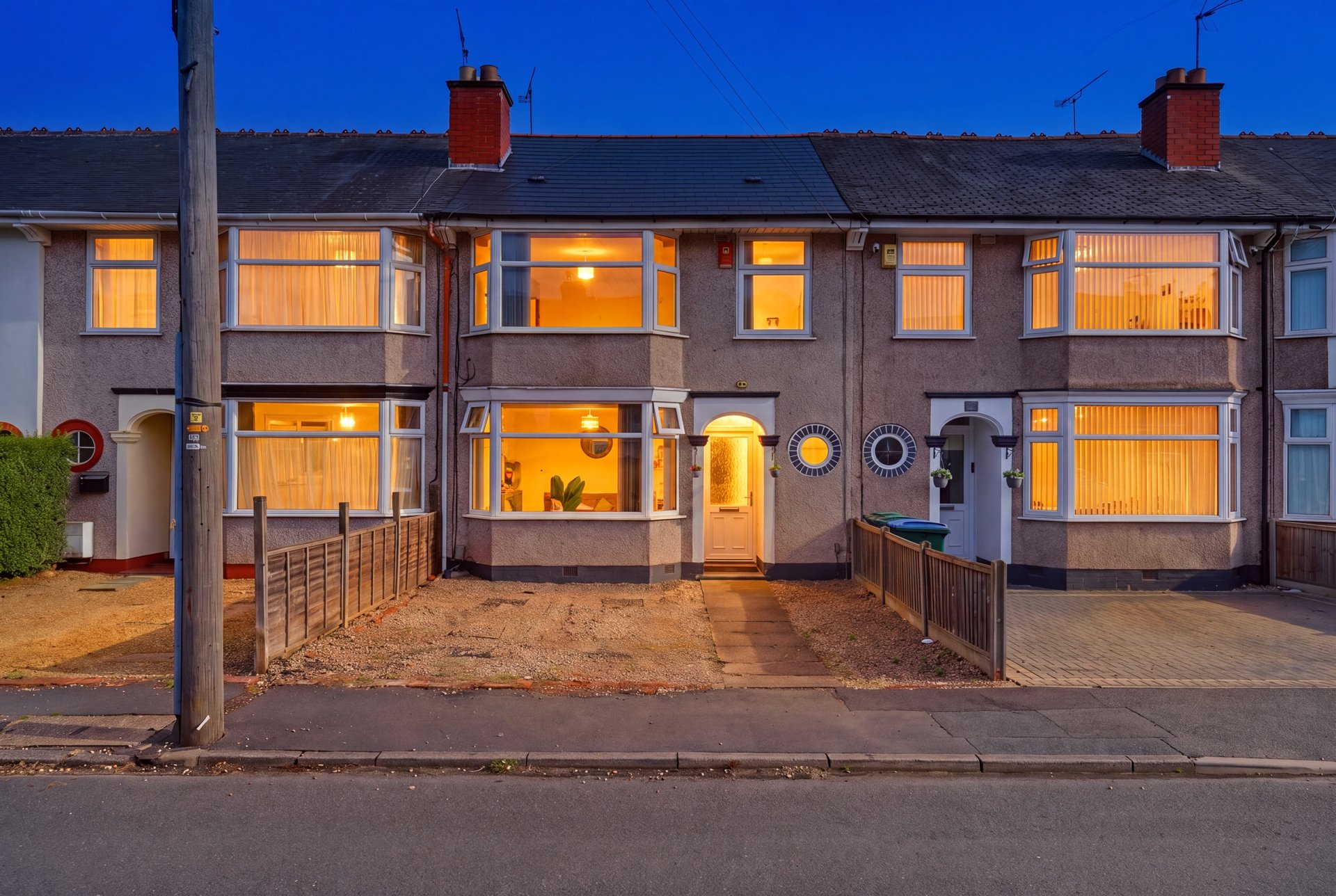 Brick terraced houses at dusk with illuminated windows, driveway and front gardens