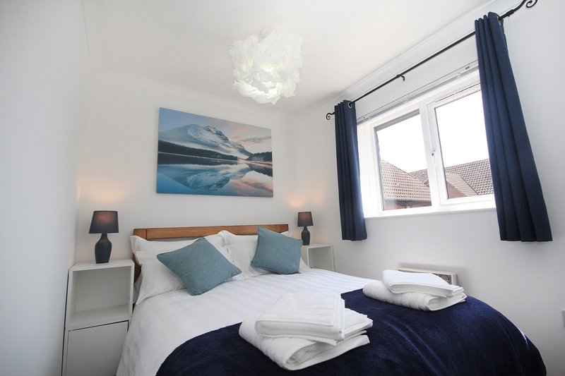 Bright bedroom with double bed, blue cushions, framed landscape artwork, bedside lamps and dark curtains