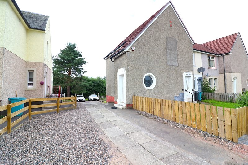 Exterior view of stone cottage with driveway, wooden fence, and adjacent residential buildings