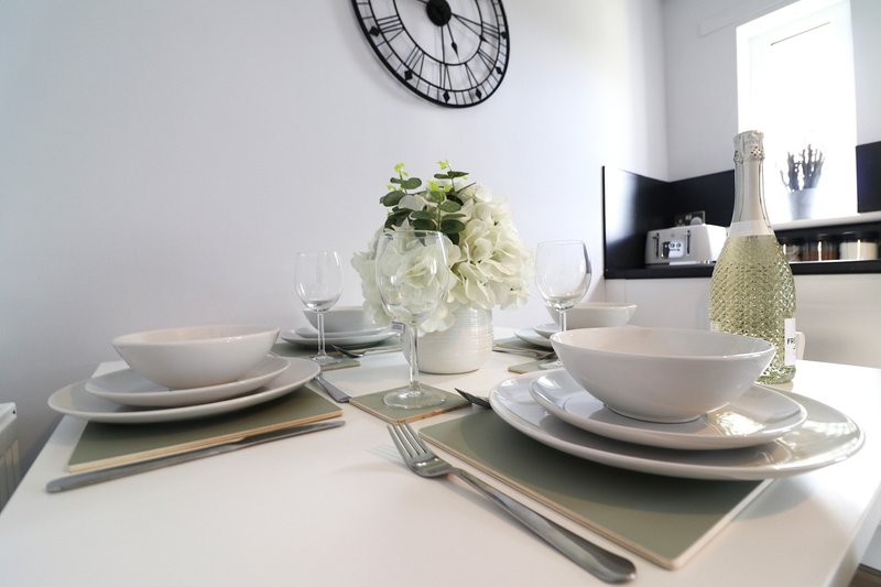 Dining table set with white plates, glasses, cutlery and floral centerpiece under wall clock