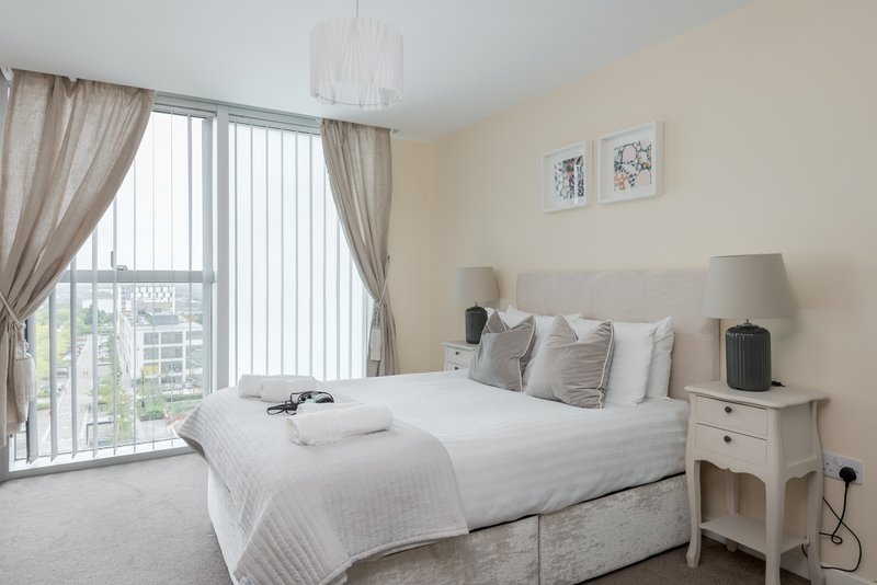 Modern bedroom with double bed, grey headboard, white bedding, and city view from floor-to-ceiling windows