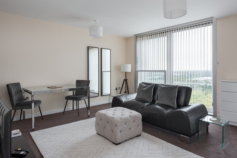Modern studio apartment with black leather sofa, desk area, and floor-to-ceiling windows overlooking cityscape