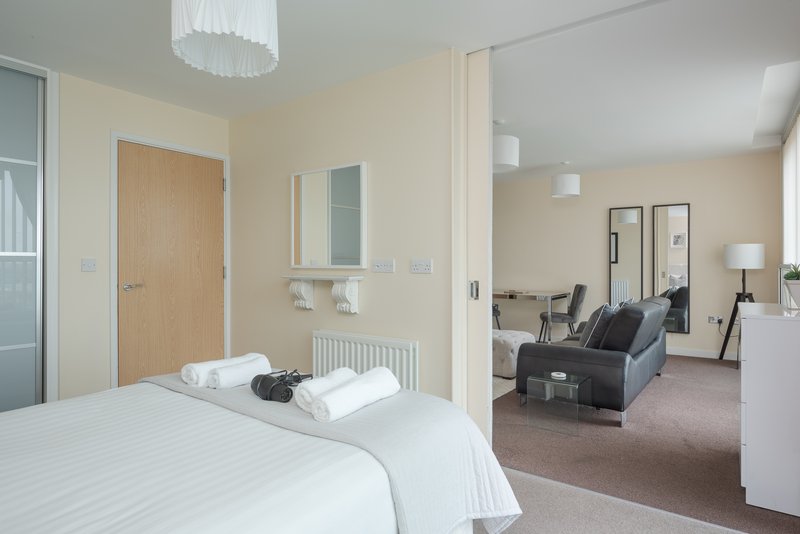 Modern bedroom with double bed, white bedding, wooden door, and open view into living area