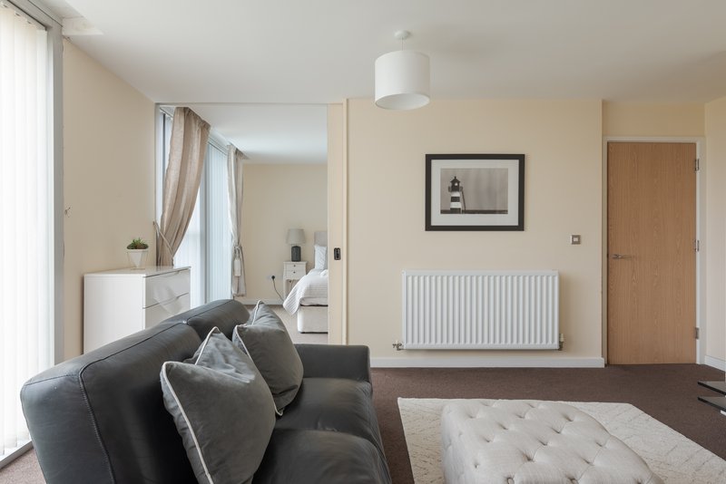 Open-plan living area with dark sofa, radiator, framed artwork, and view into bedroom
