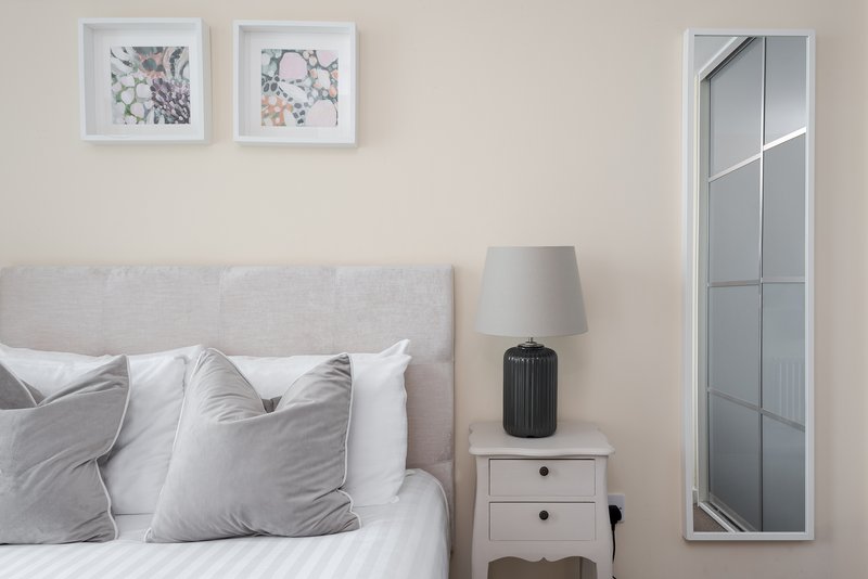 Bedroom with grey upholstered headboard, white bedding, pillows, bedside table with lamp and mirror