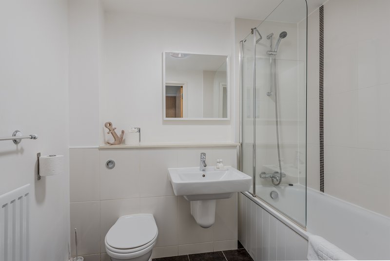 Clean bathroom with white toilet, pedestal sink, mirror, and shower bath with glass screen
