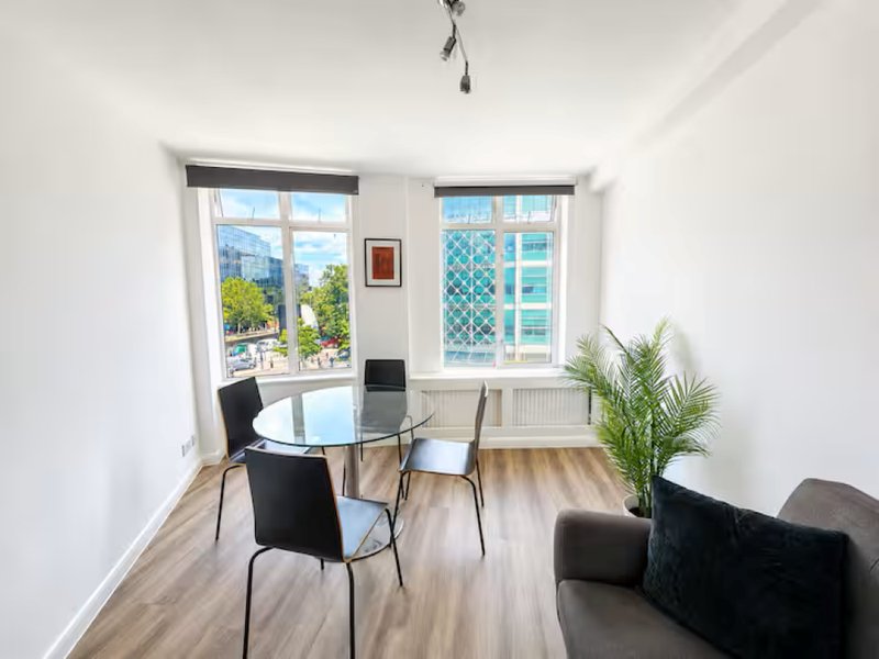 Modern living room with round glass table, black chairs, wooden flooring, and large windows overlooking city