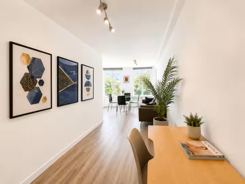 Modern hallway with wooden floor, framed wall art, desk, potted plants and open-plan living area beyond