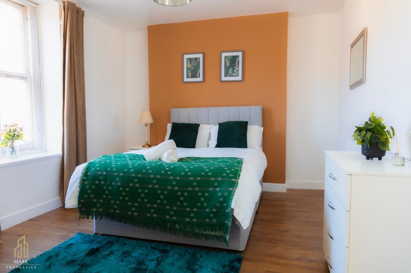 Bedroom with orange accent wall, double bed with green bedding, white dresser, and wooden flooring