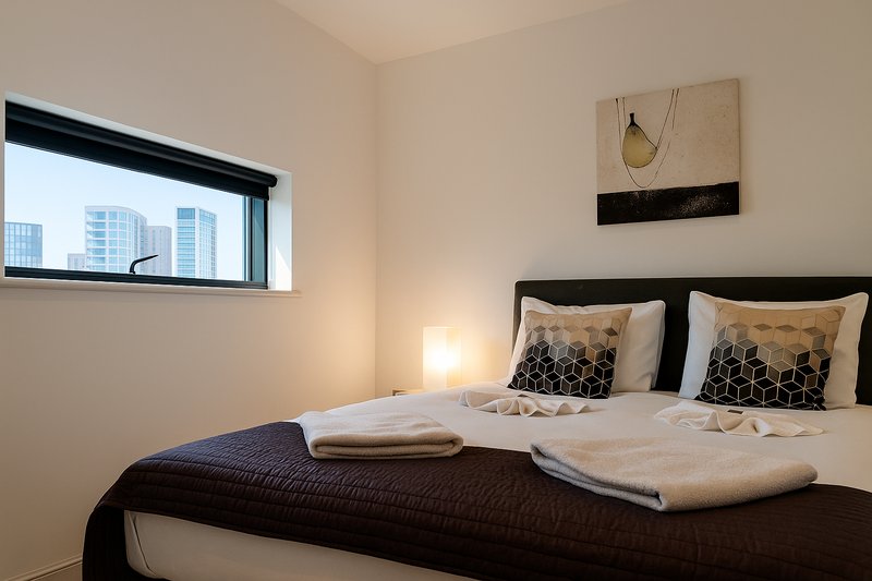 Modern bedroom with double bed, patterned cushions, folded towels, and city view window