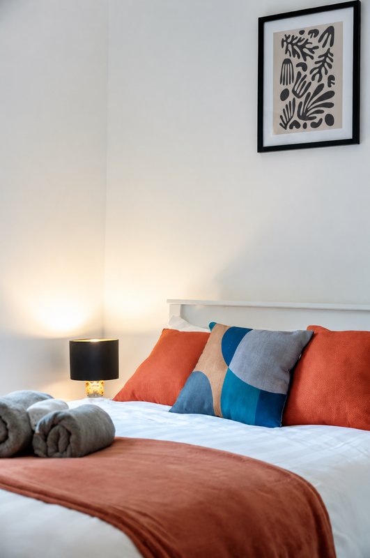 Bedroom with double bed, colorful pillows, folded towels, bedside lamp and framed wall art