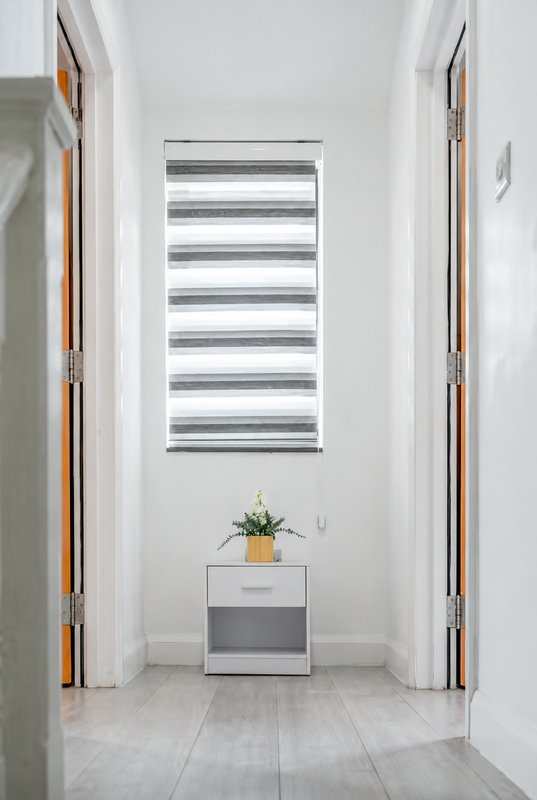 Bright hallway with striped window blind, potted plant on small cabinet, and light tile flooring