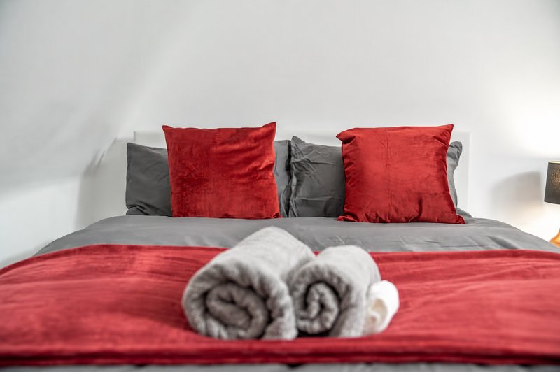 Bedroom with red and grey bedding, red velvet cushions, and rolled towels on bed