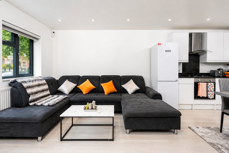 Open-plan living room with black sectional sofa, white kitchen, and large windows