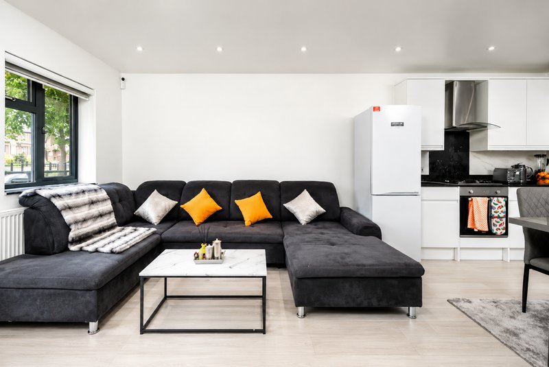 Open-plan living room with large dark grey corner sofa, white kitchen with black splashback and appliances, large windows with street view