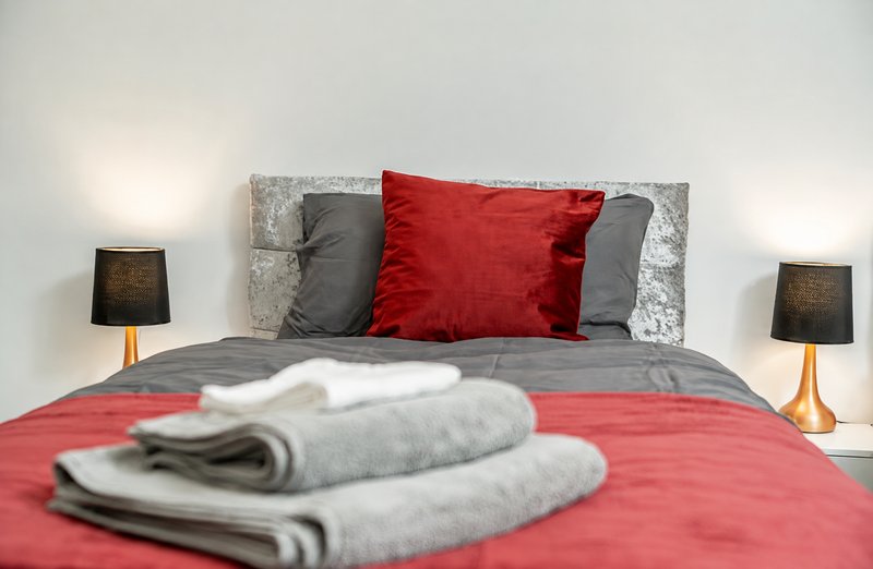 Bedroom with double bed, red bedding, gray and red pillows, folded towels, and bedside lamps