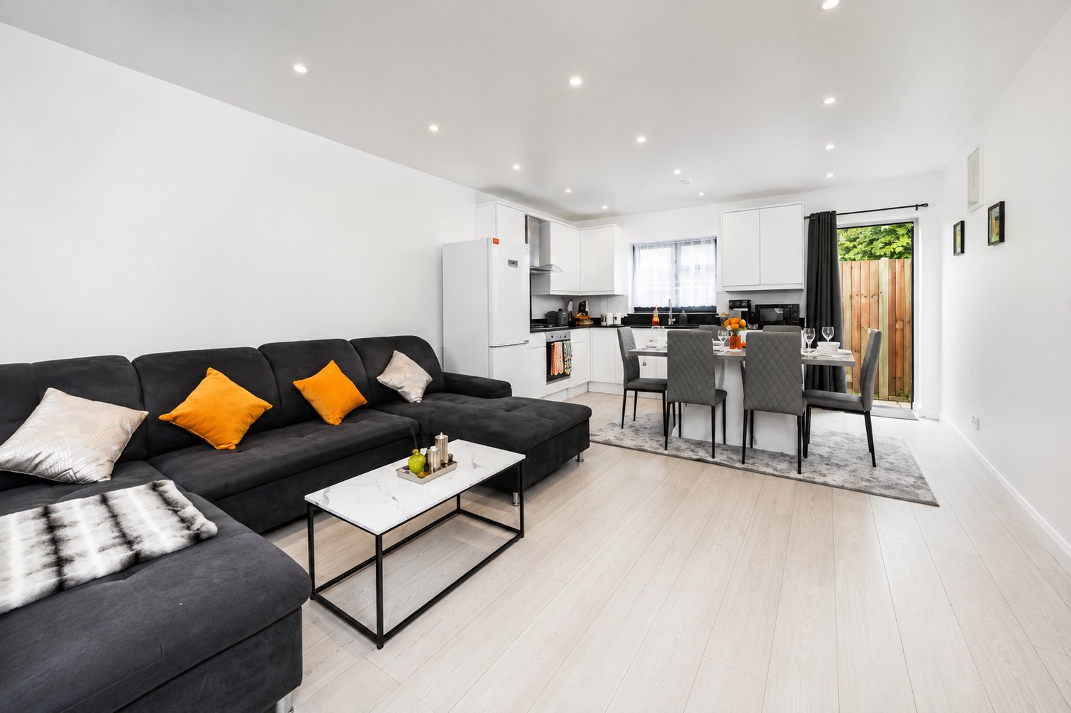 Open-plan living area with black sofa, orange cushions, dining table and kitchen visible