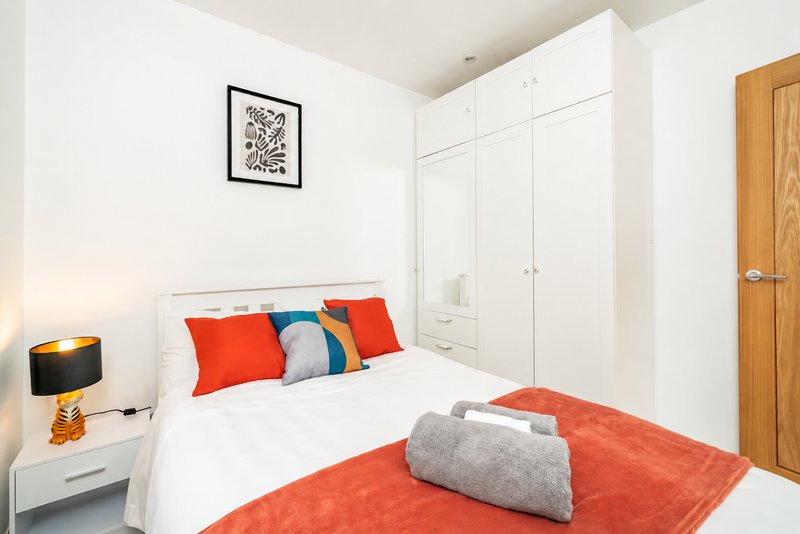 Modern bedroom with double bed, red and blue pillows, white wardrobe, wooden door and bedside lamp