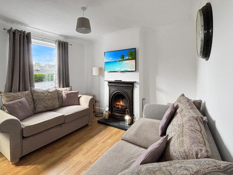 Bright living room with grey sofa, armchair, fireplace with TV above, wooden floor and large window