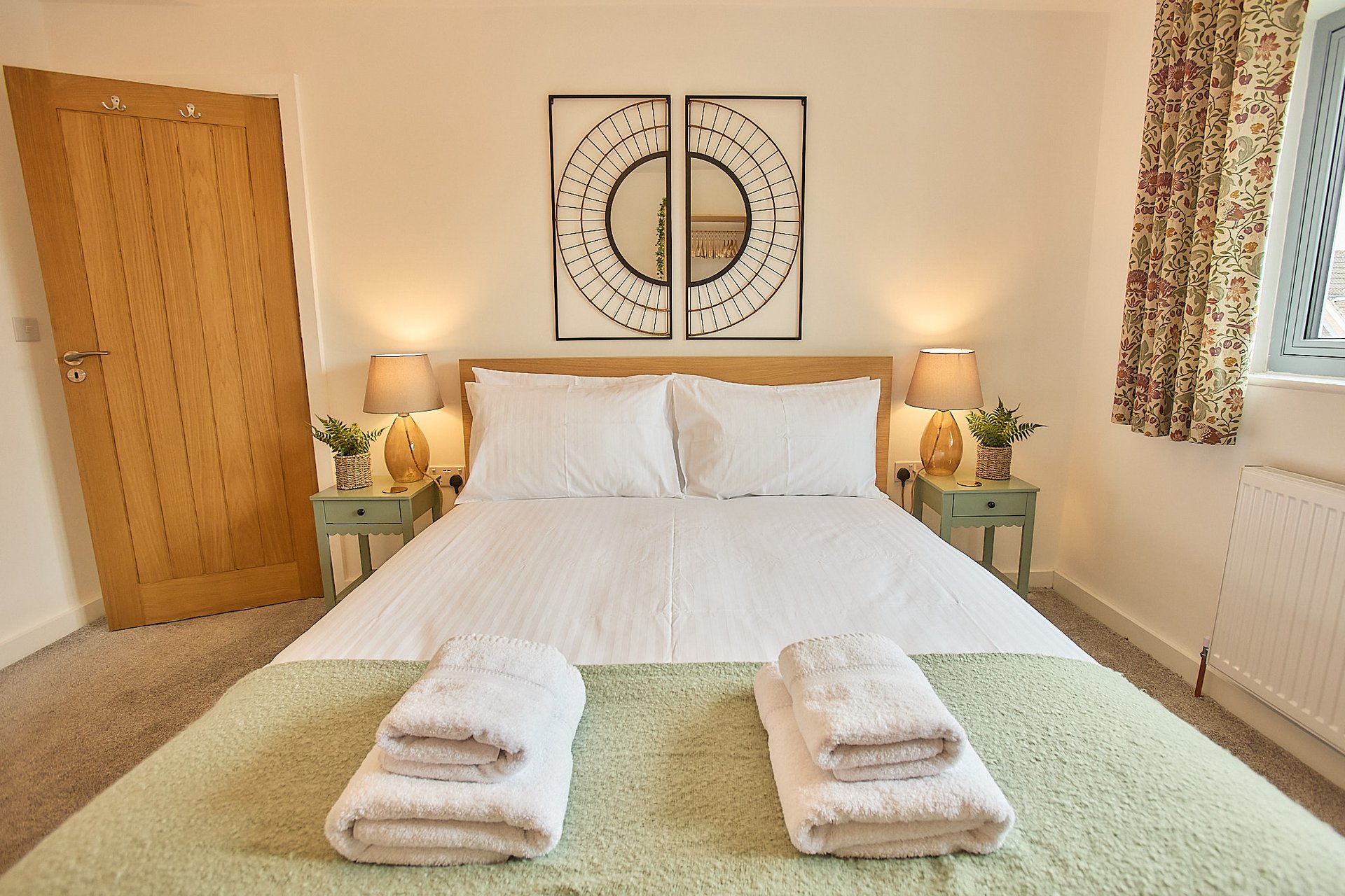 Bright bedroom with double bed, wooden door, bedside lamps, and folded towels on green throw