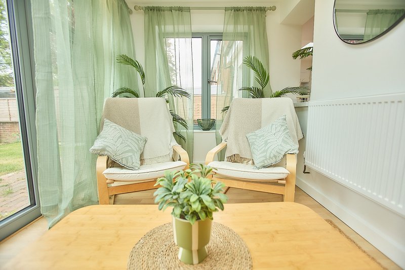Bright sunroom with wooden table, potted plants, cushioned seating and green curtains