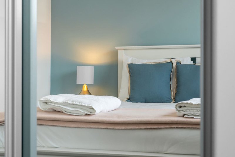 Bedroom with neatly made bed, blue cushions, bedside lamp and folded linens