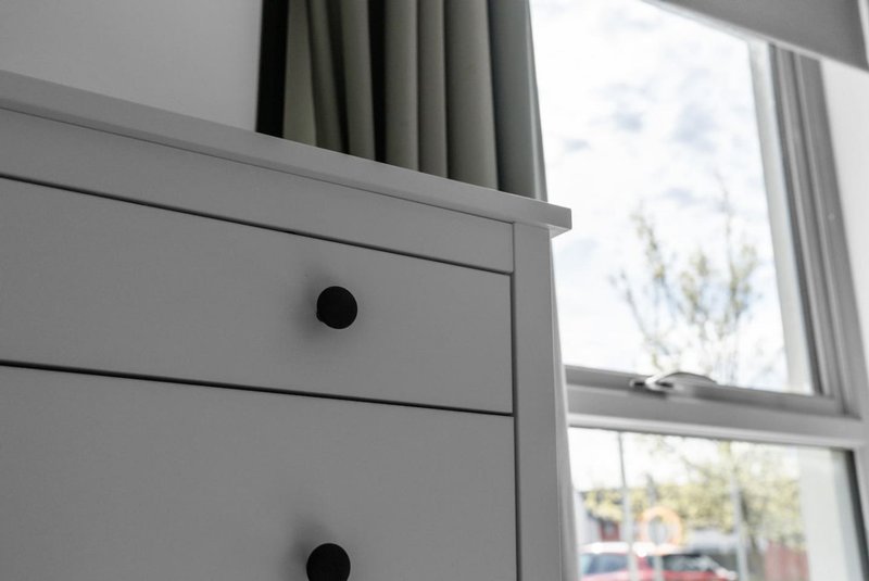 Gray chest of drawers with black knobs beside window with green curtains