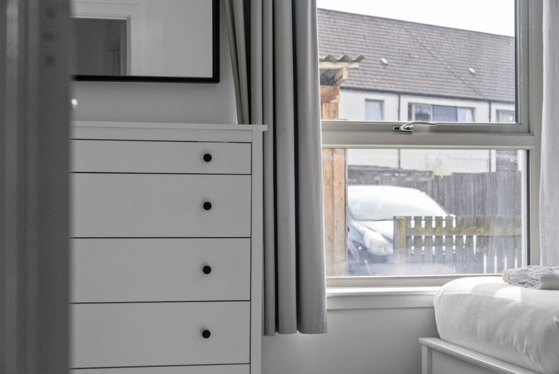 Bedroom with white dresser, window overlooking garden and neighboring houses, bed visible right side