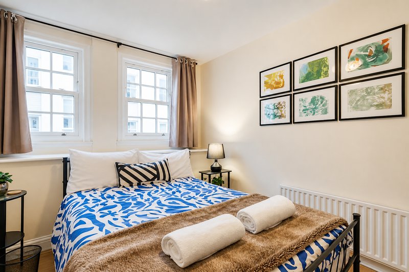 Bright bedroom with double bed, blue patterned duvet, rolled towels, framed botanical prints and windows