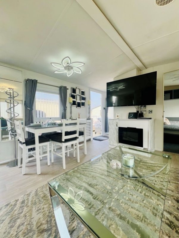 Open-plan living and dining area with white furniture, dining table, kitchen, and mounted TV