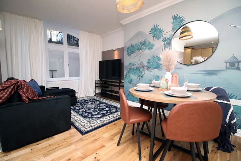 Open-plan living and dining area with black sofa, wooden dining table, teal wall mural and round mirror