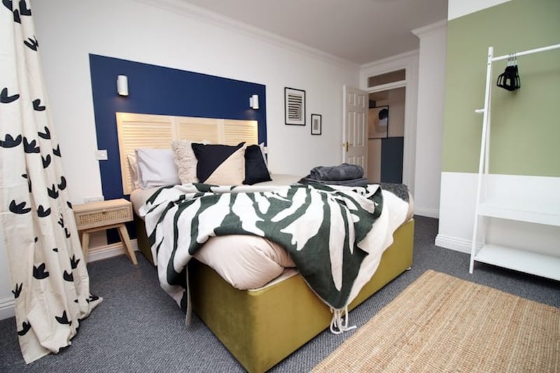 Modern bedroom with double bed, navy headboard, striped bedding, yellow footstool, and green accent wall
