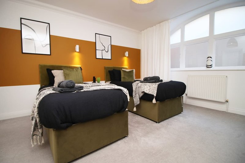 Twin bedroom with olive-green upholstered beds, black bedding, orange accent wall, and large windows.