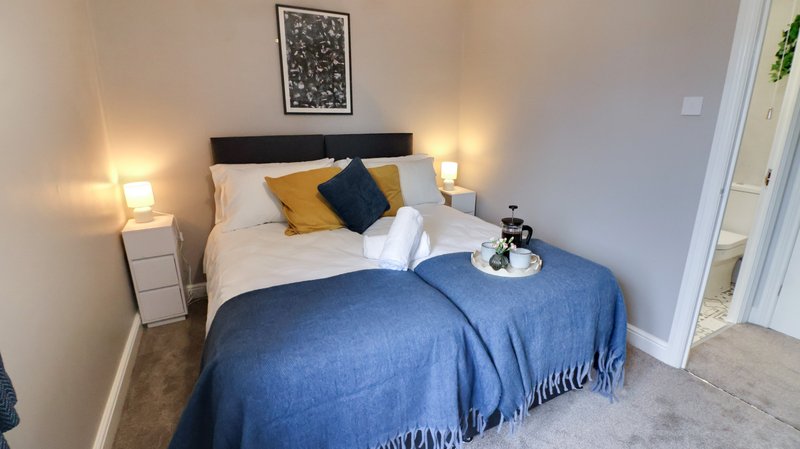 Modern bedroom with double bed, blue throw, yellow and navy cushions, bedside lamps and framed wall art